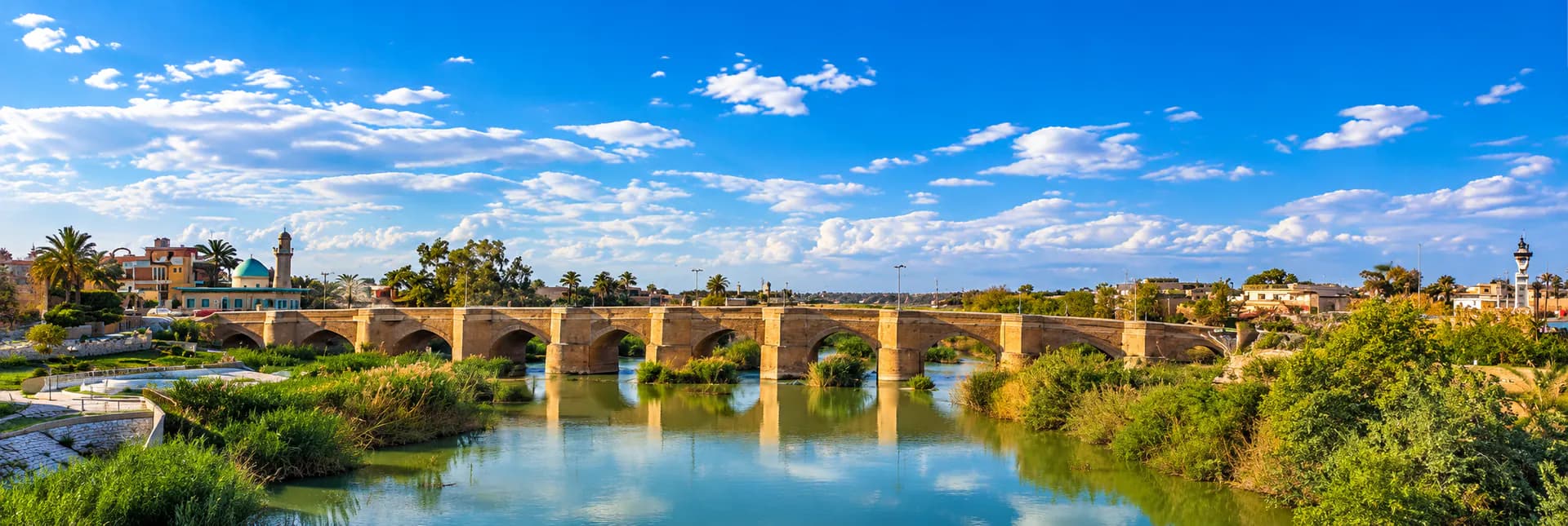 An old stone bridge with arches over a river, with greenery and palm trees around it